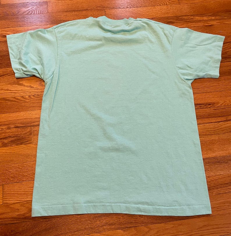 May include: A pale green, short-sleeved t-shirt. The shirt has a crew neck and is made of a soft material. The t-shirt is laid flat on a wooden surface. The shirt is a solid colour and has a simple, classic design.