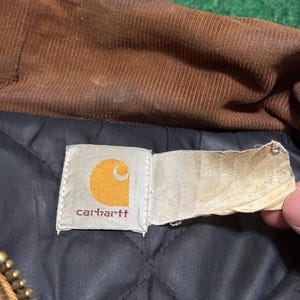 Może przedstawiać: Zbliżenie na metkę kurtki Carhartt. Etykieta jest prostokątna z białym tłem i logo Carhartt w kolorze pomarańczowym i białym. Kurtka ma brązowy kołnierz sztruksowy i czarną pikowaną podszewkę.