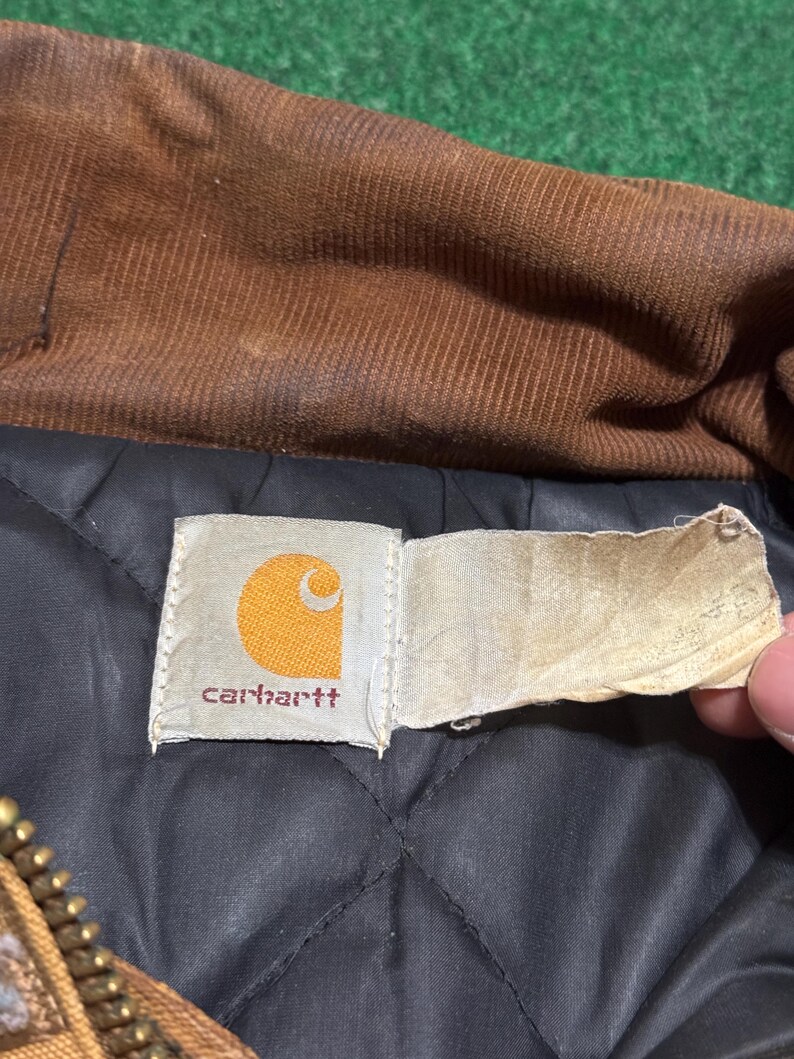 Może przedstawiać: Zbliżenie na metkę kurtki Carhartt. Etykieta jest prostokątna z białym tłem i logo Carhartt w kolorze pomarańczowym i białym. Kurtka ma brązowy kołnierz sztruksowy i czarną pikowaną podszewkę.