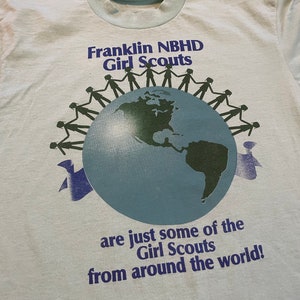 May include: Pale blue t-shirt featuring the text "Franklin NBHD Girl Scouts" and a globe graphic. The globe is encircled by silhouettes of people holding hands. The text "are just some of the Girl Scouts from around the world!" is below.