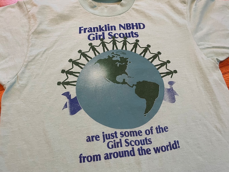 May include: Pale blue t-shirt featuring the text "Franklin NBHD Girl Scouts" and a globe graphic. The globe is encircled by silhouettes of people holding hands. The text "are just some of the Girl Scouts from around the world!" is below.