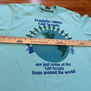 May include: A pale blue t-shirt with the text "Franklin NBHD Girl Scouts" above a globe graphic. The shirt also features the text "are just some of the Girl Scouts from around the world!" A wooden ruler is placed across the shirt.