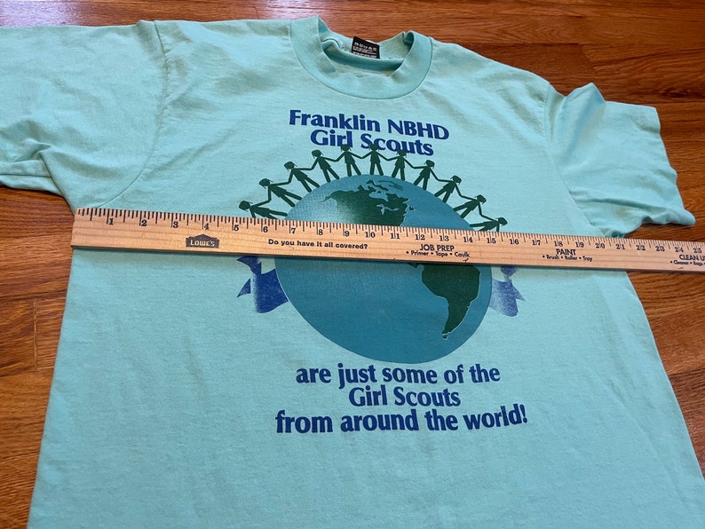 May include: A pale blue t-shirt with the text "Franklin NBHD Girl Scouts" above a globe graphic. The shirt also features the text "are just some of the Girl Scouts from around the world!" A wooden ruler is placed across the shirt.