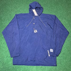 Puede incluir: Un cortavientos azul marino con capucha, cierre de un solo botón y un pequeño logotipo en la parte delantera. La chaqueta tiene capucha con cordón y cordón blanco y gris.