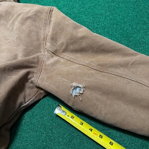 Puede incluir: Primer plano de una manga de chaqueta marr&oacute;n con un agujero. Una cinta m&eacute;trica amarilla se coloca en diagonal sobre la manga, mostrando medidas en pies y pulgadas. La chaqueta parece estar hecha de un material duradero y desgastado.