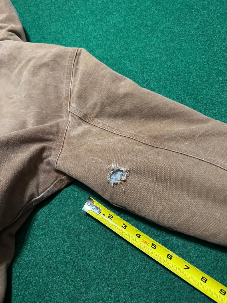 Puede incluir: Primer plano de una manga de chaqueta marr&oacute;n con un agujero. Una cinta m&eacute;trica amarilla se coloca en diagonal sobre la manga, mostrando medidas en pies y pulgadas. La chaqueta parece estar hecha de un material duradero y desgastado.