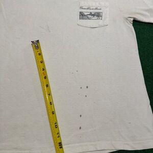May include: A white short-sleeve t-shirt with a chest pocket featuring a black graphic of a shark and the text Ron Jon Surf Shop One of a Kind Cocoa Beach Florida. A yellow measuring tape shows a length of approximately 14 inches on the shirt.