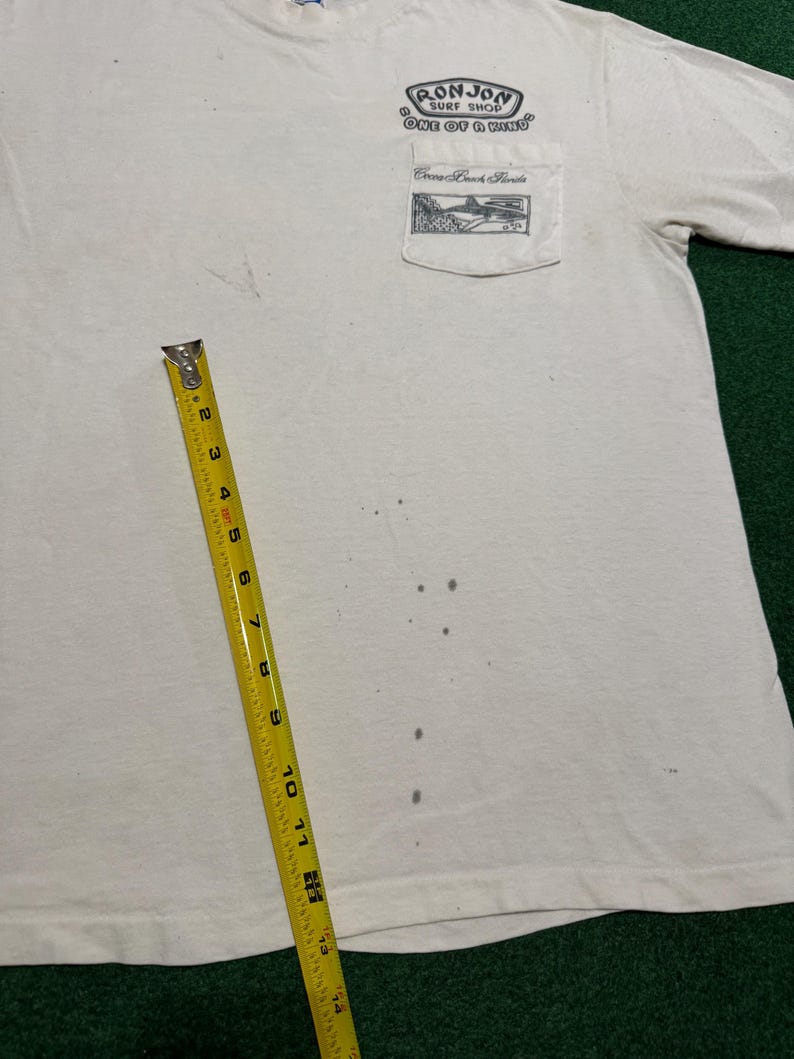 May include: A white short-sleeve t-shirt with a chest pocket featuring a black graphic of a shark and the text Ron Jon Surf Shop One of a Kind Cocoa Beach Florida. A yellow measuring tape shows a length of approximately 14 inches on the shirt.