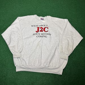 Pode incluir: Um moletom cinza claro com o texto "WHAT ABOUT... J2C JESUS' SECOND COMING" em preto e vermelho. O moletom tem mangas compridas e gola redonda.
