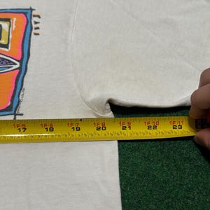May include: A white t-shirt featuring a colorful graphic print with blue, orange, and purple accents. A yellow measuring tape is positioned across the chest, showing a width measurement extending from 17 inches to 24 inches across the garment.
