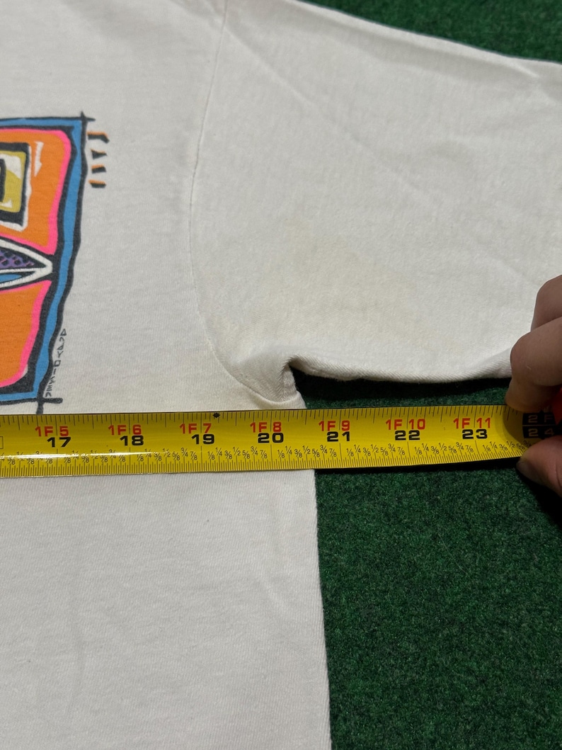 May include: A white t-shirt featuring a colorful graphic print with blue, orange, and purple accents. A yellow measuring tape is positioned across the chest, showing a width measurement extending from 17 inches to 24 inches across the garment.