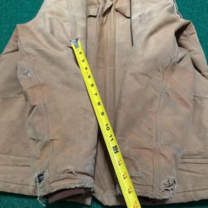 Może przedstawiać: Zużyta, beżowa kurtka robocza z brązowym kołnierzem i ż&oacute;łtą miarką. Kurtka nosi ślady użytkowania, w tym postrzępione krawędzie i małe dziury. Miarka wskazuje długość około 46 cm.