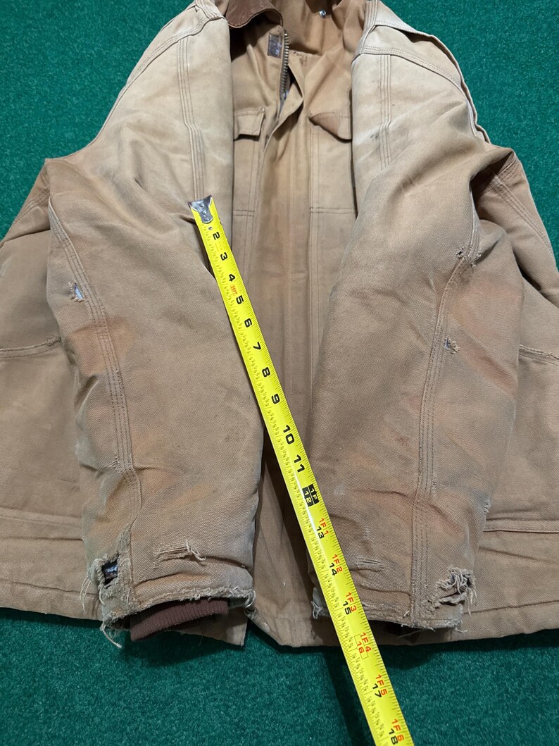 Może przedstawiać: Zużyta, beżowa kurtka robocza z brązowym kołnierzem i ż&oacute;łtą miarką. Kurtka nosi ślady użytkowania, w tym postrzępione krawędzie i małe dziury. Miarka wskazuje długość około 46 cm.