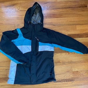 Puede incluir: Una chaqueta de esquí negra, blanca y azul con capucha. La chaqueta tiene una cremallera y dos bolsillos en la parte delantera.