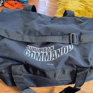 Könnte beinhalten: Schwarze Reisetasche mit einem weißen "SUBURBAN COMMANDO" Logo auf der Vorderseite. Die Tasche hat einen schwarzen Reißverschluss und einen schwarzen Riemen.