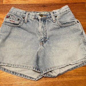 May include: Light blue denim shorts with a faded wash and a small red logo on the left pocket. The shorts have a high waist and a relaxed fit.