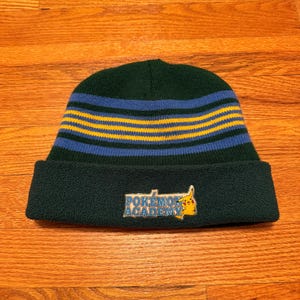 May include: A dark green beanie hat with blue and yellow horizontal stripes. The folded-over brim displays a rectangular patch with the text "Pokemon Academy" and a Pikachu graphic.