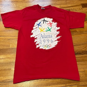 Puede incluir: Una camiseta roja con el logotipo de los Juegos Olímpicos de Atlanta 1996. El logotipo incluye los anillos olímpicos, el texto "Atlanta 1996" y un diseño estilizado con anillos y estrellas de colores. La camiseta es de material suave.