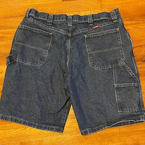 Puede incluir: Pantalones cortos de trabajo de mezclilla azul oscuro con múltiples bolsillos y una etiqueta de logotipo roja.