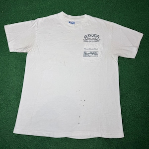 May include: A white short-sleeve t-shirt with a chest pocket featuring a black graphic print that reads Ron Jon Surf Shop One of a Kind and Cocoa Beach Florida. The shirt has a crew neckline and several small dark spots on the front fabric.