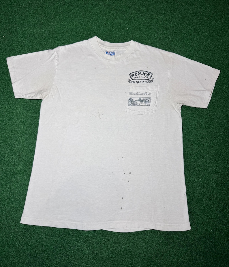 May include: A white short-sleeve t-shirt with a chest pocket featuring a black graphic print that reads Ron Jon Surf Shop One of a Kind and Cocoa Beach Florida. The shirt has a crew neckline and several small dark spots on the front fabric.