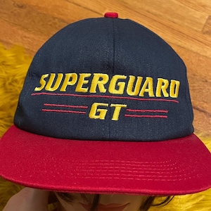 May include: A navy blue and red baseball cap with the text "SUPERGUARD GT" embroidered in yellow.