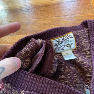 May include: A brown and beige patterned sweater with a V-neck and a zipper. The sweater has a label that reads "Heirloom Collectibles".