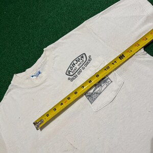 May include: A white short-sleeve t-shirt with a chest pocket featuring a black graphic of a surfboard and the text Ron Jon Surf Shop One of a Kind. A yellow measuring tape is placed across the shirt, showing a width of approximately 15 inches.