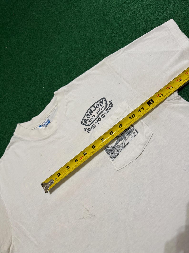 May include: A white short-sleeve t-shirt with a chest pocket featuring a black graphic of a surfboard and the text Ron Jon Surf Shop One of a Kind. A yellow measuring tape is placed across the shirt, showing a width of approximately 15 inches.