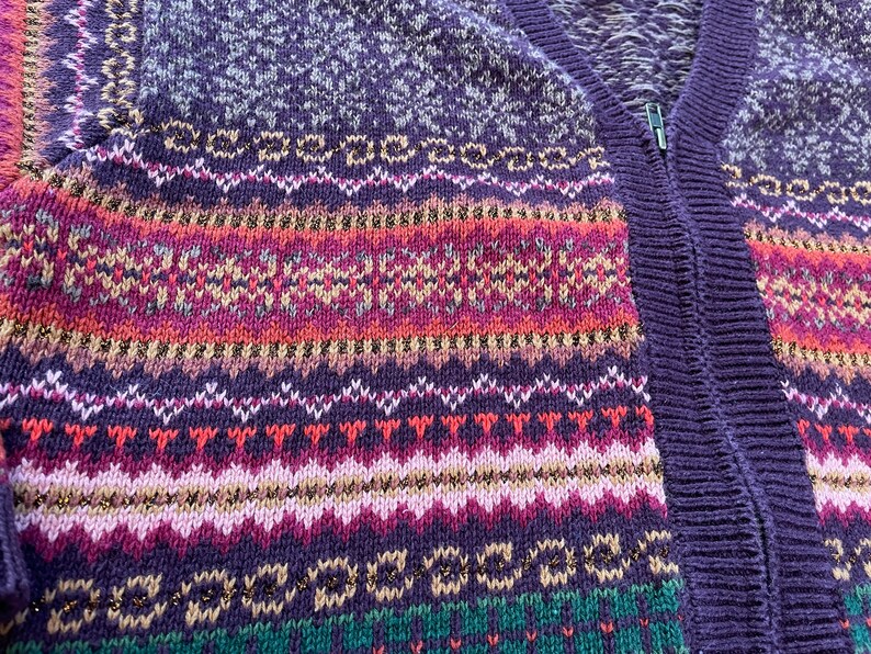 May include: Purple, pink, green, and white knitted cardigan with a zipper closure. The cardigan has a fair isle pattern with gold thread accents.