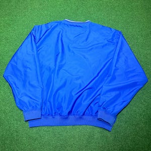 以下が含まれることがあります： 白の襟トリムと青のリブ編みの裾と袖口が付いた、鮮やかな青色のウィンドブレーカー。 ジャケットは軽量素材で作られています。 ジャケットは緑色の表面に表示されています。