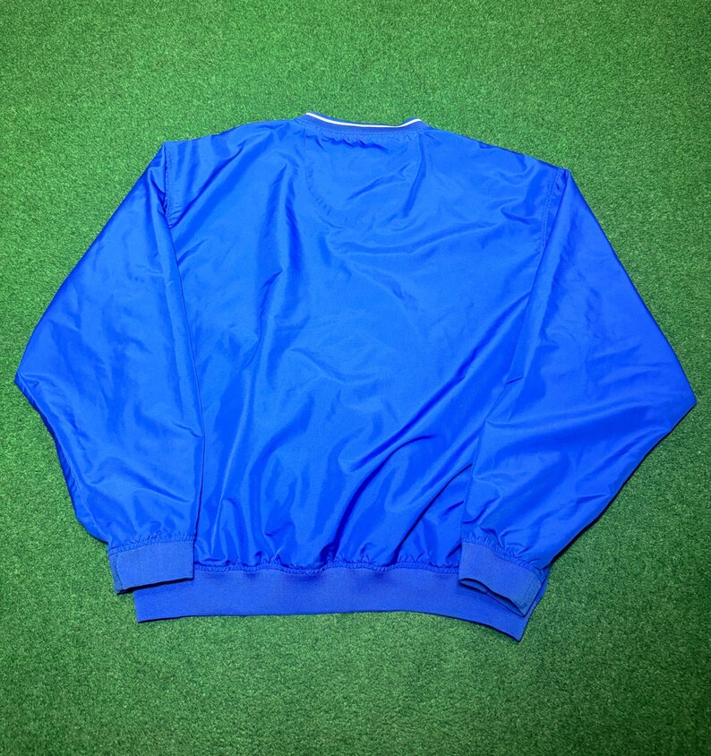 以下が含まれることがあります： 白の襟トリムと青のリブ編みの裾と袖口が付いた、鮮やかな青色のウィンドブレーカー。 ジャケットは軽量素材で作られています。 ジャケットは緑色の表面に表示されています。