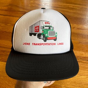May include: A white trucker hat with a black mesh back. The front of the hat features a green and red illustration of a semi-truck with a trailer. The text "LYONS TRANSPORTATION LINES" is printed in black below the truck.
