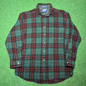 Puede incluir: Una camisa abotonada a cuadros verdes, rojos y negros. La camisa de manga larga tiene cuello y puños acolchados. La camisa está abotonada y colocada sobre una superficie verde.