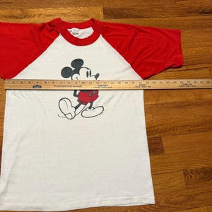 Pu&ograve; includere: T-shirt bianco e rosso a maniche raglan con grafica di Topolino. La grafica mostra Topolino in nero, rosso e bianco. La maglietta misura circa 71 cm di lunghezza.
