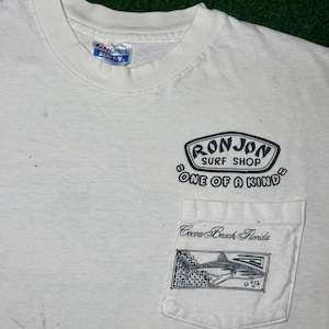 May include: A white short-sleeved t-shirt with a crew neck and a chest pocket. The shirt features black printed text that reads Ron Jon Surf Shop One of a Kind and Cocoa Beach Florida, along with a graphic illustration of a shark on the pocket.