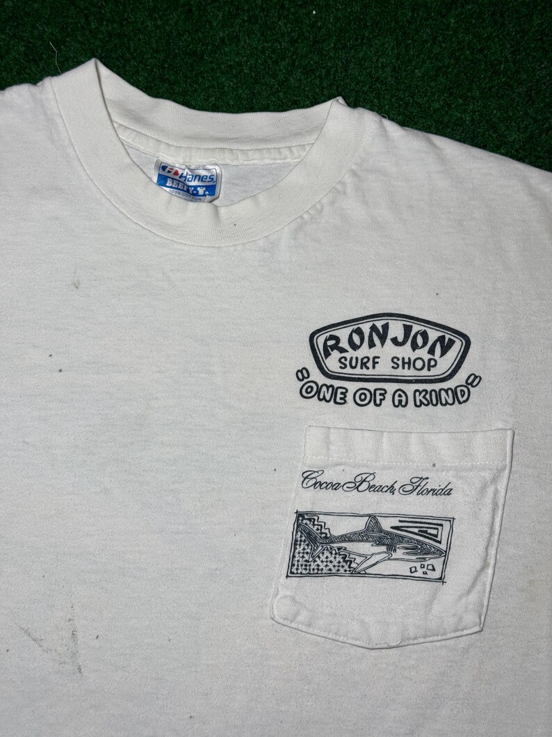 May include: A white short-sleeved t-shirt with a crew neck and a chest pocket. The shirt features black printed text that reads Ron Jon Surf Shop One of a Kind and Cocoa Beach Florida, along with a graphic illustration of a shark on the pocket.