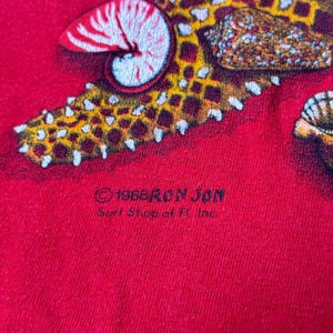 May include: Red t-shirt with a starfish and seashell design. The shirt has the text "1988 Ron Jon Surf Shop of FL Inc." printed on the bottom.