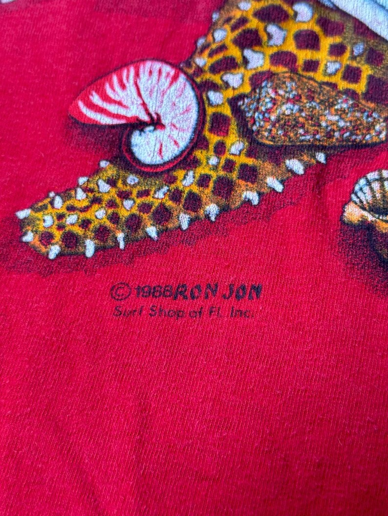 May include: Red t-shirt with a starfish and seashell design. The shirt has the text "1988 Ron Jon Surf Shop of FL Inc." printed on the bottom.