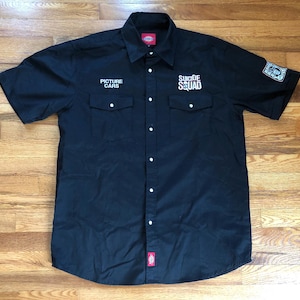 May include: A black short-sleeved button-down shirt with the text "Suicide Squad" and "Picture Cars" embroidered on the front.