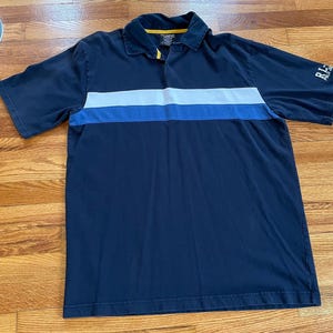 Puede incluir: Polo azul marino con una franja horizontal blanca y azul en el pecho. La camisa tiene mangas cortas y cuello. La marca "RL POLO" es visible en la manga.