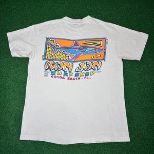 May include: A white short-sleeve t-shirt featuring a graphic on the back. The design includes a blue shark inside an orange rectangular frame with yellow accents. Below the shark, the text reads RON JON SURF SHOP COCOA BEACH, FL in stylized letters.