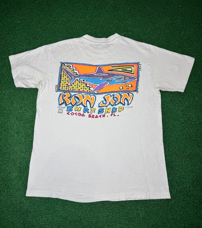 May include: A white short-sleeve t-shirt featuring a graphic on the back. The design includes a blue shark inside an orange rectangular frame with yellow accents. Below the shark, the text reads RON JON SURF SHOP COCOA BEACH, FL in stylized letters.