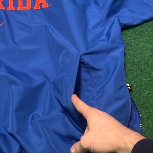 以下が含まれることがあります： オレンジ色の文字で「RIDA」と書かれた青いナイロンジャケット。 ジャケットにはジッパー開閉とポケットが付いています。 ジャケットは緑色の表面にあります。