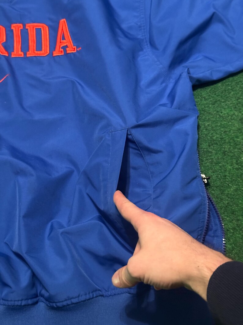 以下が含まれることがあります： オレンジ色の文字で「RIDA」と書かれた青いナイロンジャケット。 ジャケットにはジッパー開閉とポケットが付いています。 ジャケットは緑色の表面にあります。