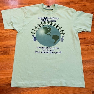 May include: A pale green t-shirt featuring the text "Franklin NBHD Girl Scouts" above a globe graphic. The globe is encircled by stick figures holding hands. The text "are just some of the Girl Scouts from around the world!" is below.