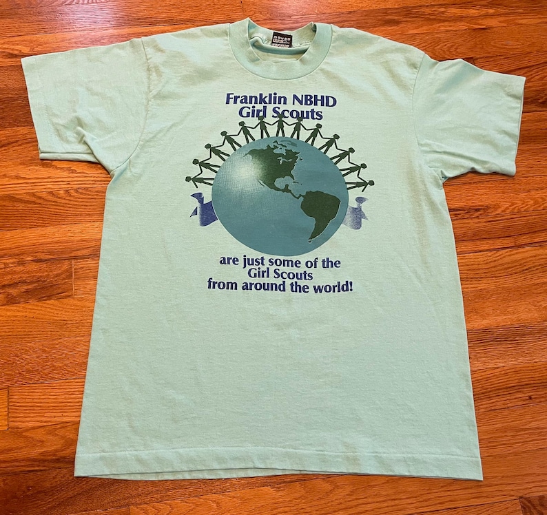 May include: A pale green t-shirt featuring the text "Franklin NBHD Girl Scouts" above a globe graphic. The globe is encircled by stick figures holding hands. The text "are just some of the Girl Scouts from around the world!" is below.