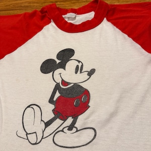Pu&ograve; includere: T-shirt bianco con maniche e colletto rossi con una grafica di Topolino sorridente. Il personaggio dei cartoni animati &egrave; raffigurato in nero, rosso e bianco. La maglietta &egrave; probabilmente in stile vintage o retr&ograve;.