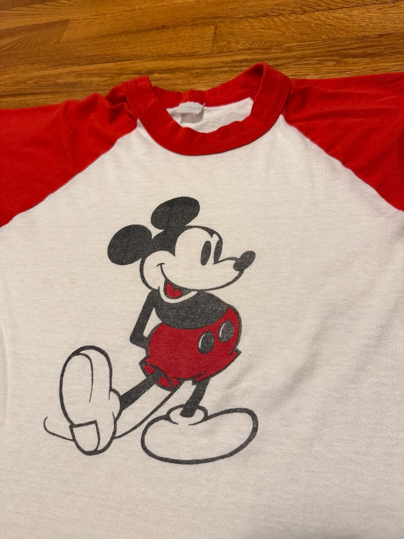 Pu&ograve; includere: T-shirt bianco con maniche e colletto rossi con una grafica di Topolino sorridente. Il personaggio dei cartoni animati &egrave; raffigurato in nero, rosso e bianco. La maglietta &egrave; probabilmente in stile vintage o retr&ograve;.