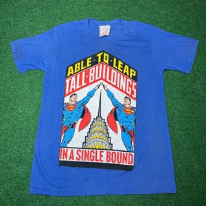 Puede incluir: Camiseta azul con un gráfico de Superman. El gráfico incluye el texto "ABLE TO LEAP TALL BUILDINGS IN A SINGLE BOUND" con ilustraciones de Superman y un rascacielos.